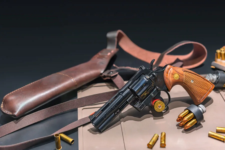 An image of a Colt Python .357 Magnum revolver with a blued finish, positioned on a table next to a leather shoulder holster. The revolver has wood grips with the Colt emblem, and it is partially loaded with .357 Magnum cartridges. Additional loose cartridges and a speed loader are arranged neatly on the table, suggesting readiness for use or display. The holster and revolver hint at a classic and professional setup, likely appealing to firearm enthusiasts and collectors.