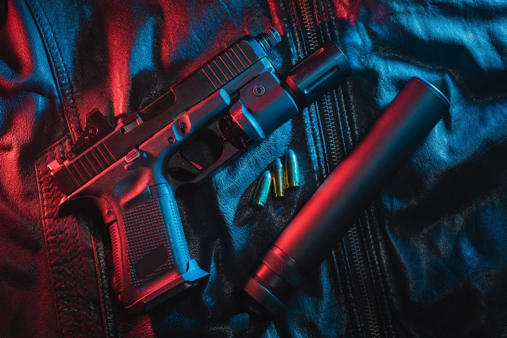 Handgun with a mounted flashlight and suppressor, placed on a dark leather surface with bullets nearby, illuminated by red and blue lighting.