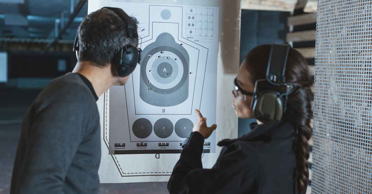 Shooting instructor pointing at a target with shot groupings at an indoor shooting range, discussing shot grouping improvement with a student. Both are wearing hearing protection and safety glasses, highlighting firearm accuracy training and shot placement correction.