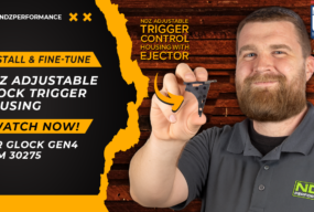 NDZ Performance team member holding an NDZ adjustable Glock trigger housing with ejector. Text reads "Install & Fine-Tune NDZ Adjustable Glock Trigger Housing – Watch Now! For Glock Gen4 9mm 30275." Includes NDZ branding, Made in USA badge, and a wood-textured background.