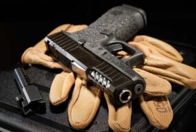 Custom Glock pistol with ported slide and stippled grip, displayed on tan shooting gloves alongside a match-grade barrel, showcasing premium aftermarket parts and build craftsmanship.