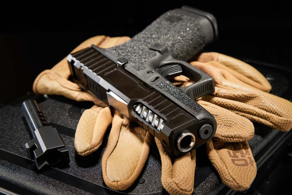Custom Glock pistol with ported slide and stippled grip, displayed on tan shooting gloves alongside a match-grade barrel, showcasing premium aftermarket parts and build craftsmanship.