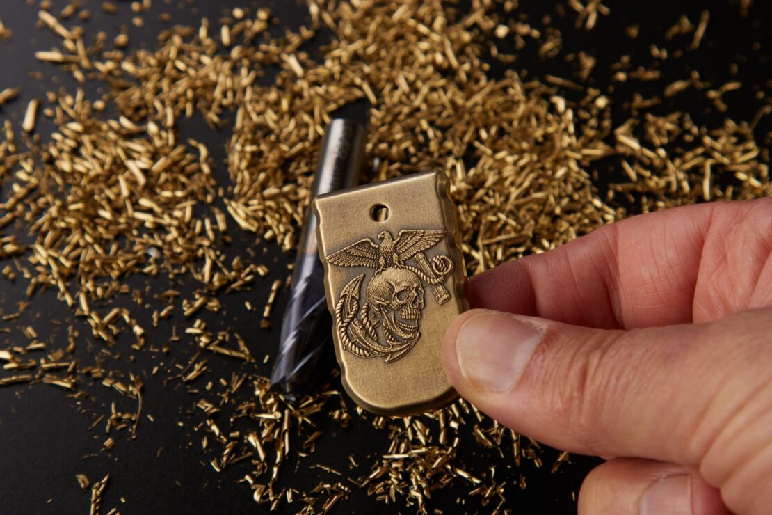 Hand holding a brass 3D magazine plate engraved with the Marine Corps emblem, over brass shavings