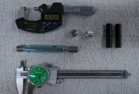 Shop measuring tools on a towel: a digital micrometer, a dial caliper, a thread plug gauge, and small machined parts